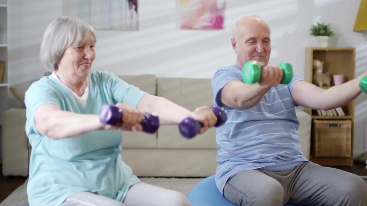 Cheerful senior man and woman sitting on exercise balls and doing dumbbell wrist twist while working out together at home